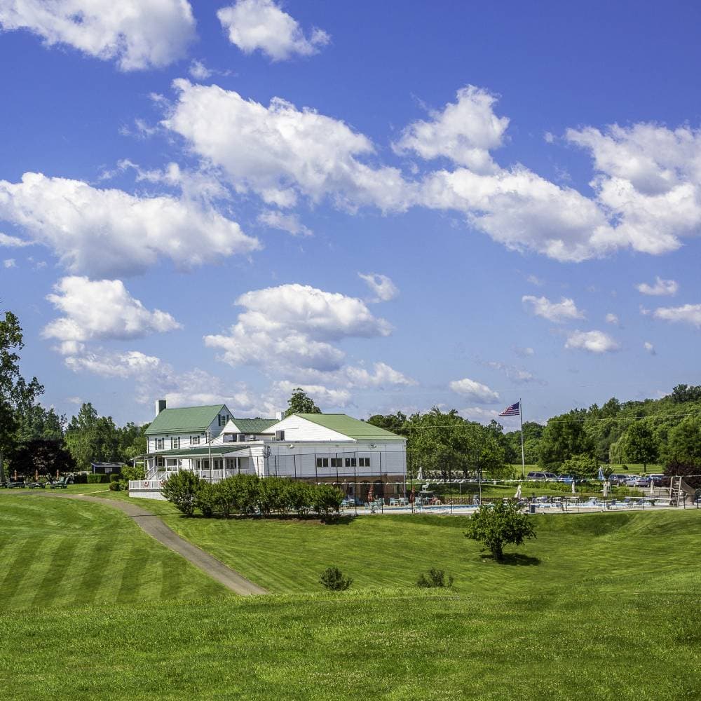 The Greene Hills clubhouse on a summer afternoon, white siding and green metal roof with the American flag and a summer sky.