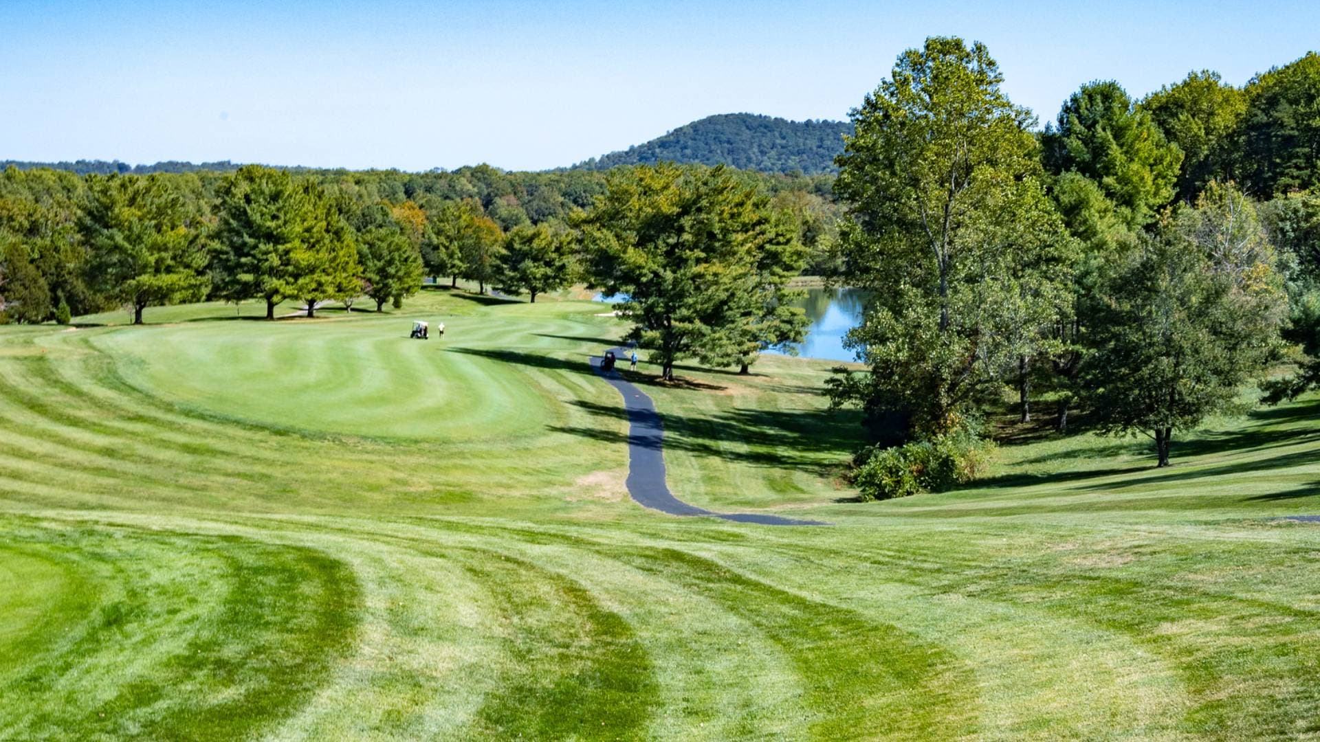 A Greene Hills fairway sweeping toward a pond and the tree-lined far bank, with the Blue Ridge in the distance.