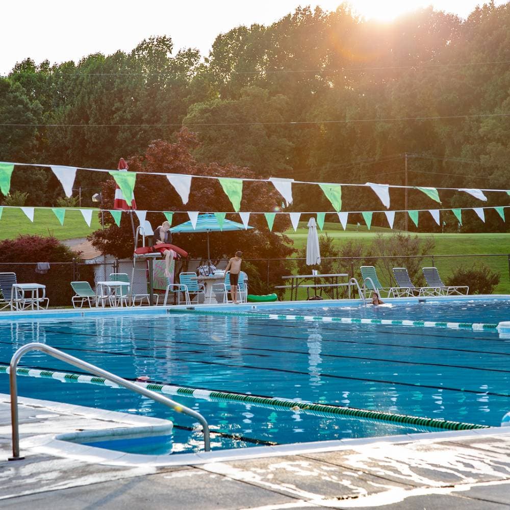 The Greene Hills pool deck at sunset with green-and-white pennants, lifeguard chairs and lens flare from the western light.